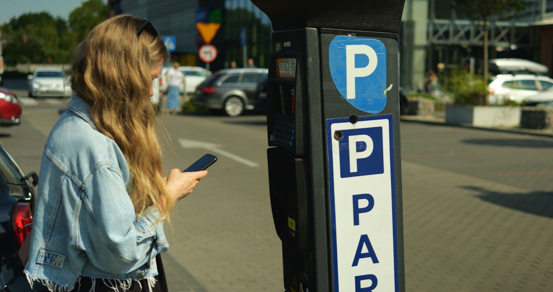 Young beautiful woman in denim jacket stand at parking meter with smartphone. Female student with long brown hair prepares to pay contactless with mobile phone at outdoors car parking lot on sunny day